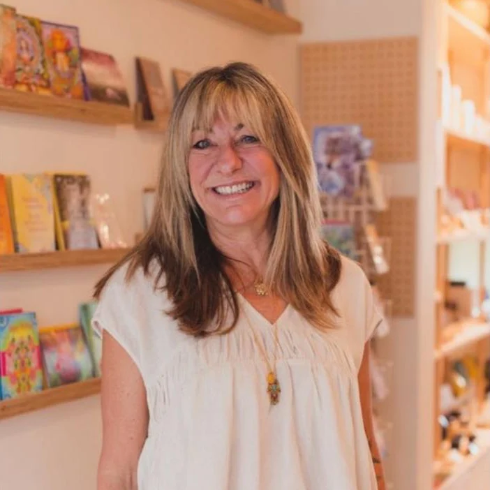 Joanne smiling in a wellbeing shop, in a cream linen top, with shelves of books and cards behind her.